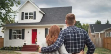 A couple with their arms around each other stands facing a two-story white house with a red door and a small yard. A couple with their arms around each other stands admiring a home they just purchased - two-story white house with a red door and a small yard.