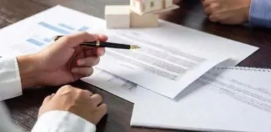 Two people at a table with documents, a small house model, and a pen in hand, suggesting a discussion or negotiation. Two people at a table with documents, a small house model, and a pen in hand, suggesting a discussion or negotiation.