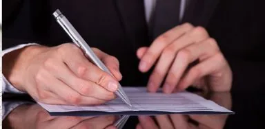 Person in a suit signing a document with a silver pen on a reflective surface. Person in a suit signing a document with a silver pen on a reflective surface.